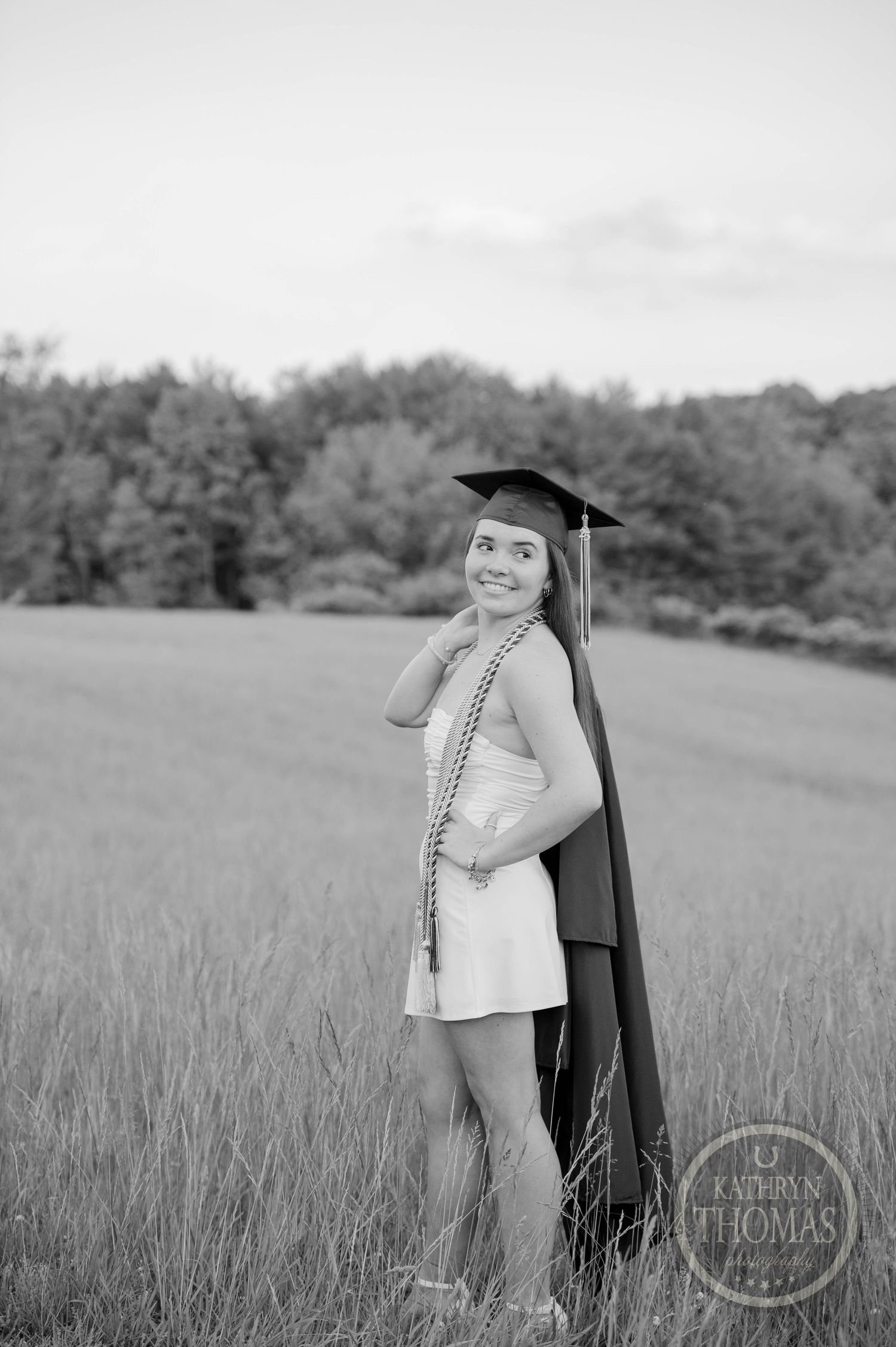 Black and white graduation portrait of someone in a cap and gown standing in a grassy field with trees in background.