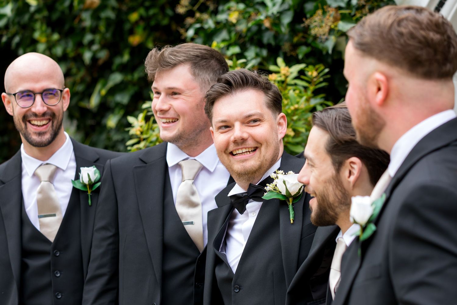 Groom laughing with his groomsmen in black suits and white rose buttonholes before the ceremony at The Bush Hotel, Farnham