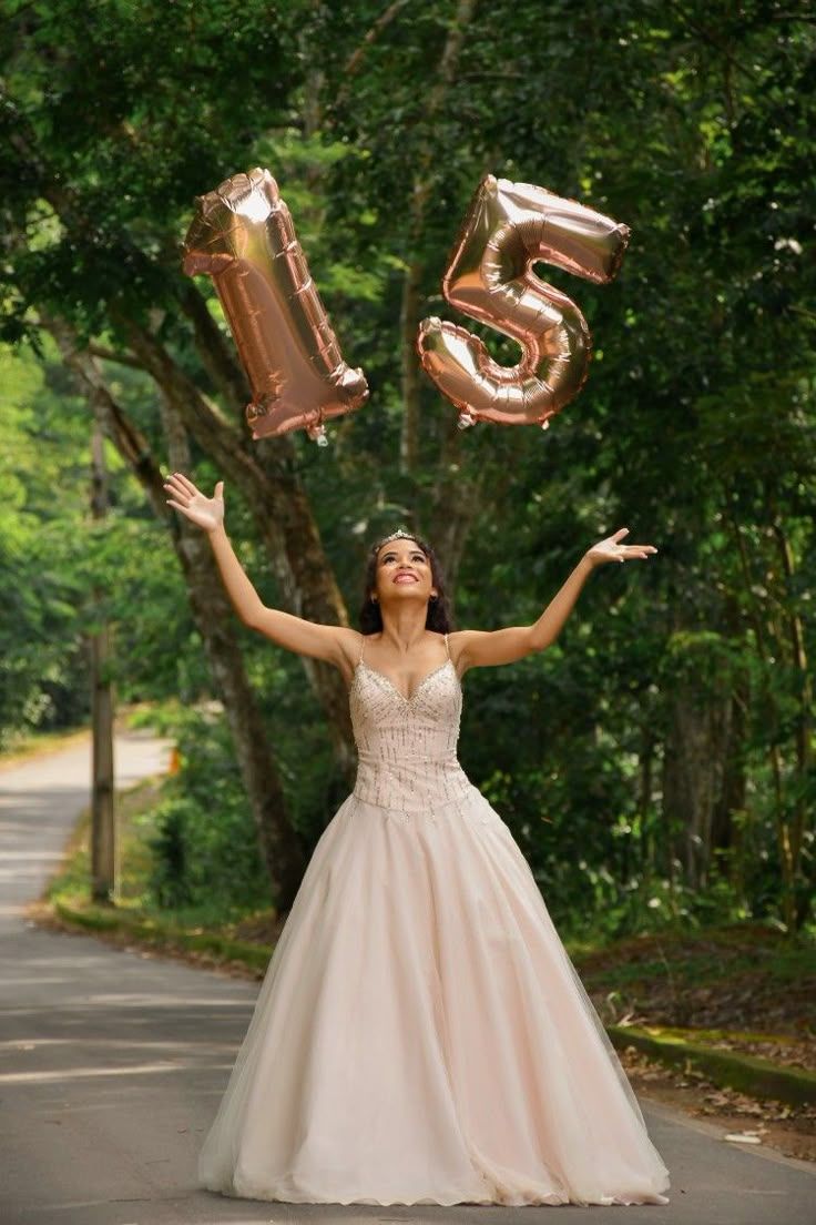 A person in a blush pink ball gown celebrates with rose gold number 15 balloons on a tree-lined path.
