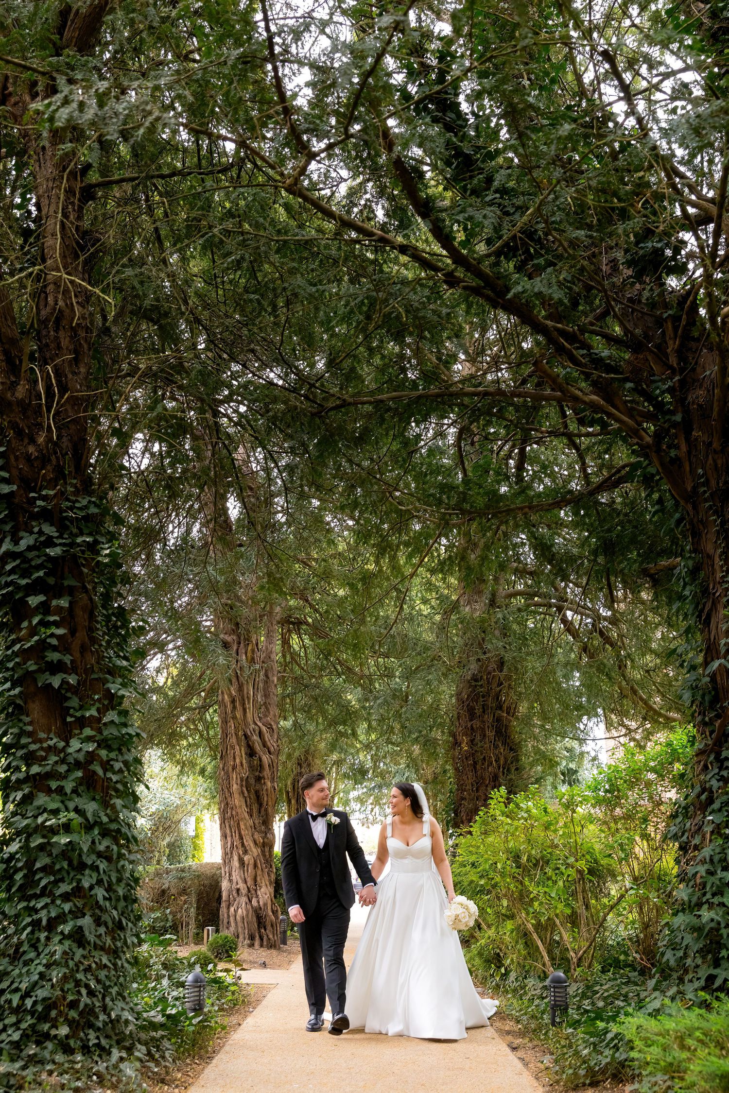 Bride and groom walking hand in hand through tree-lined path at The Bush Hotel, surrounded by lush greenery.