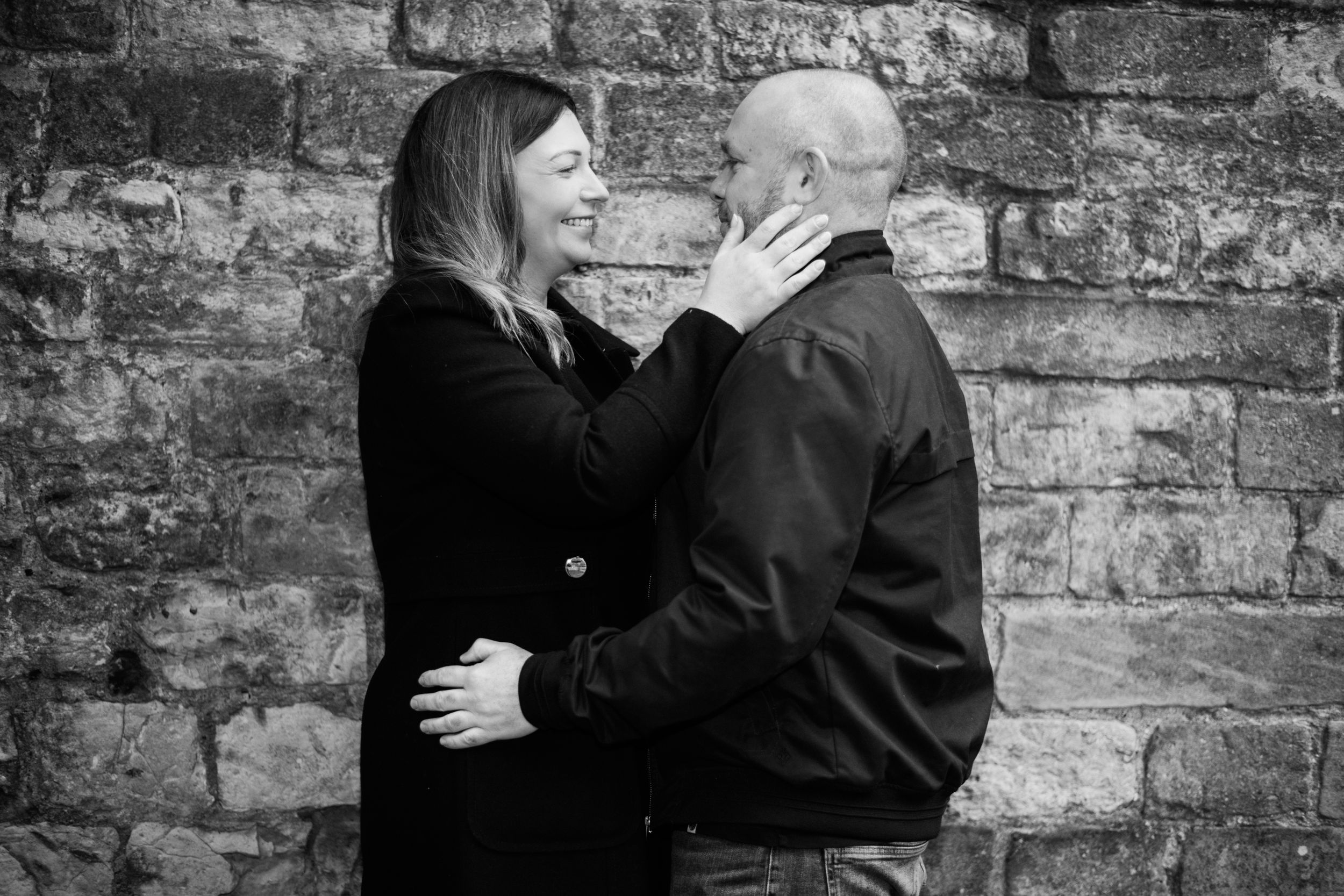 Black and white portrait of couple embracing against rustic stone wall background.
