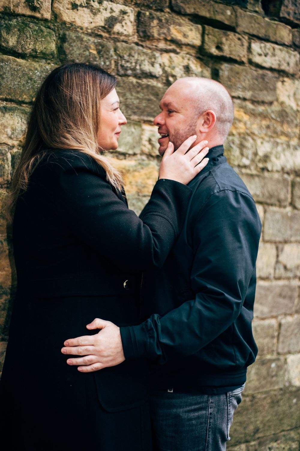 Romantic couple embracing against stone wall in intimate outdoor portrait session.
