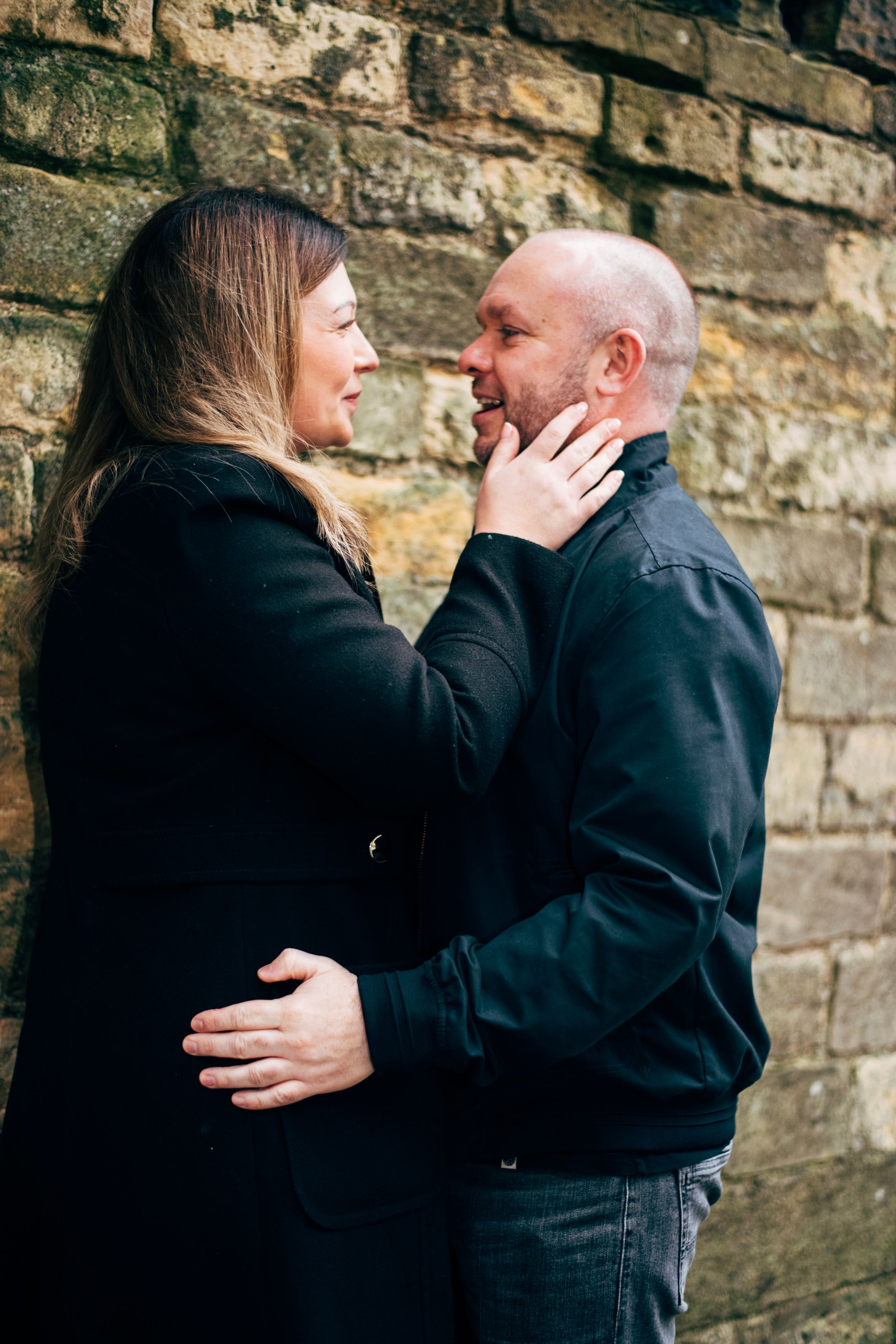 Romantic couple embracing against stone wall in intimate outdoor portrait session.