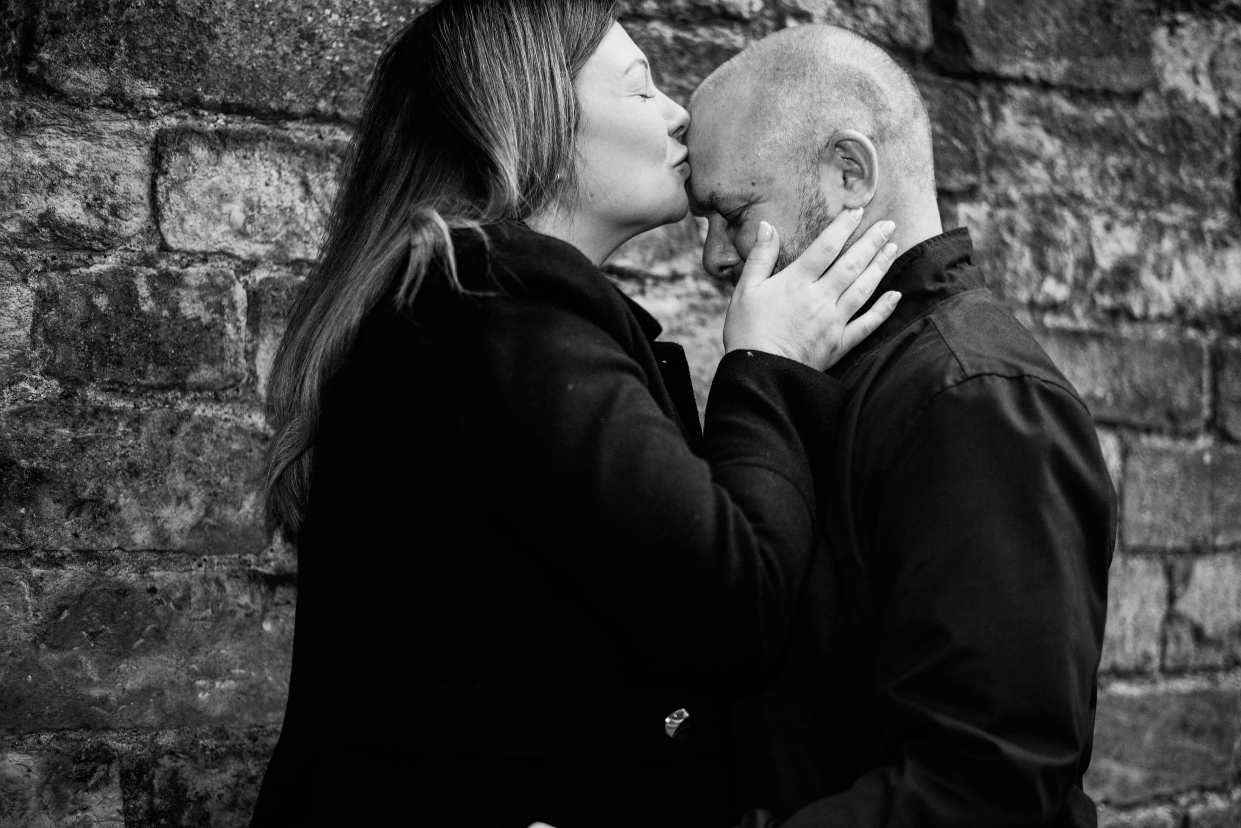 Intimate black and white portrait of couple sharing tender moment against brick wall.