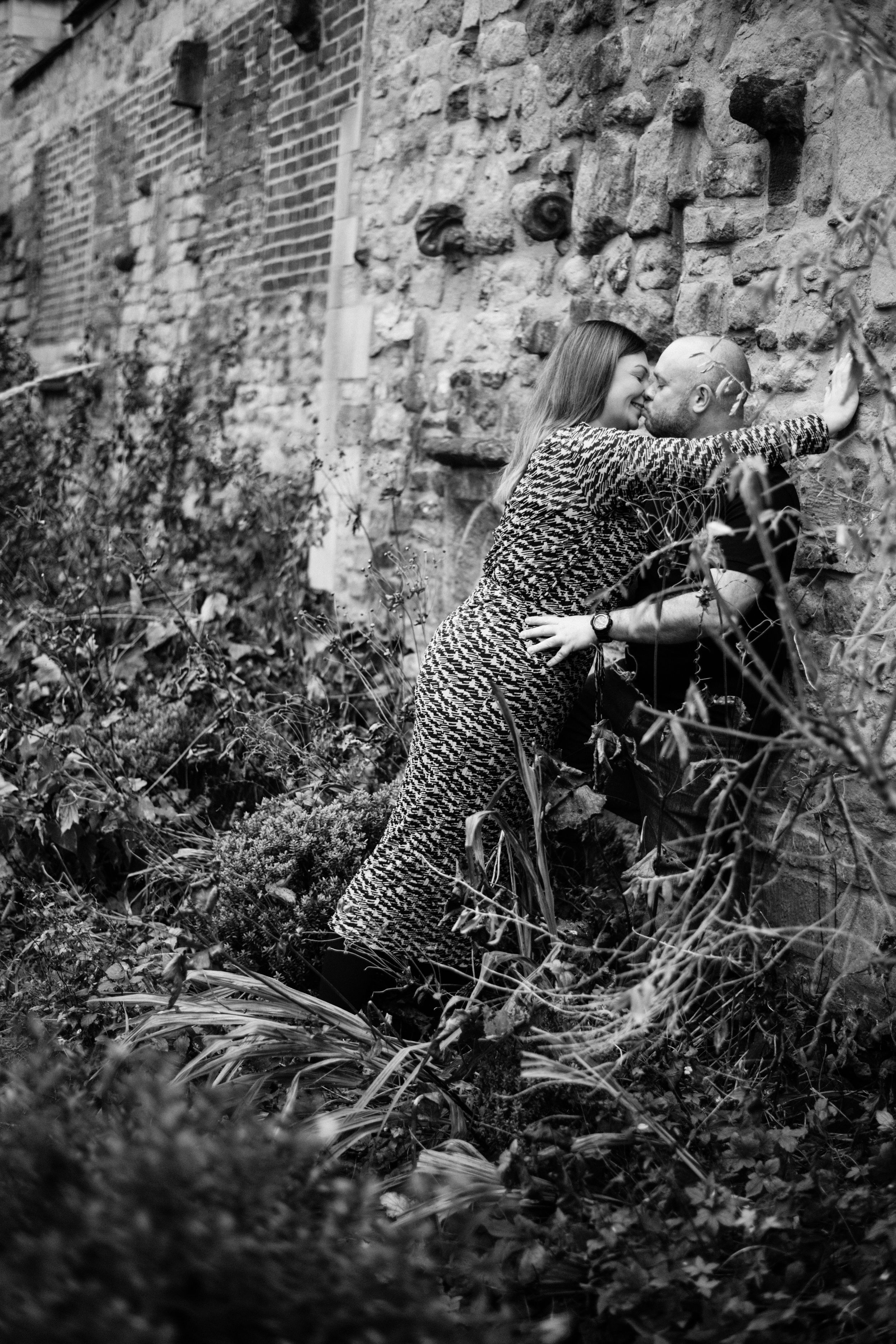 Black and white artistic photo of couple in playful pose by weathered stone wall.