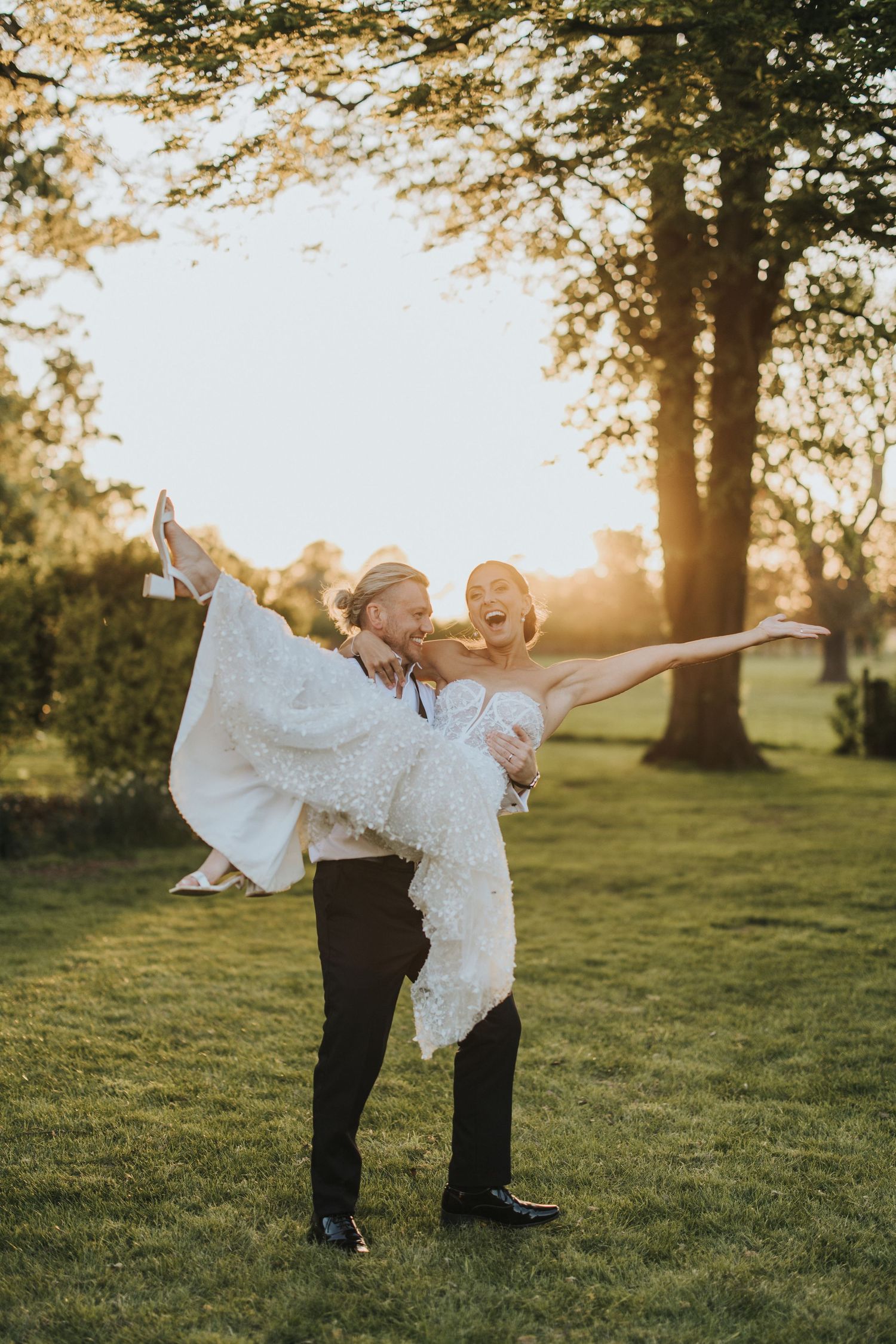 Bride with arms open is smiling in the groom’s arms as they embrace one another during a golden sunset. Setting is The Grange Leverton.