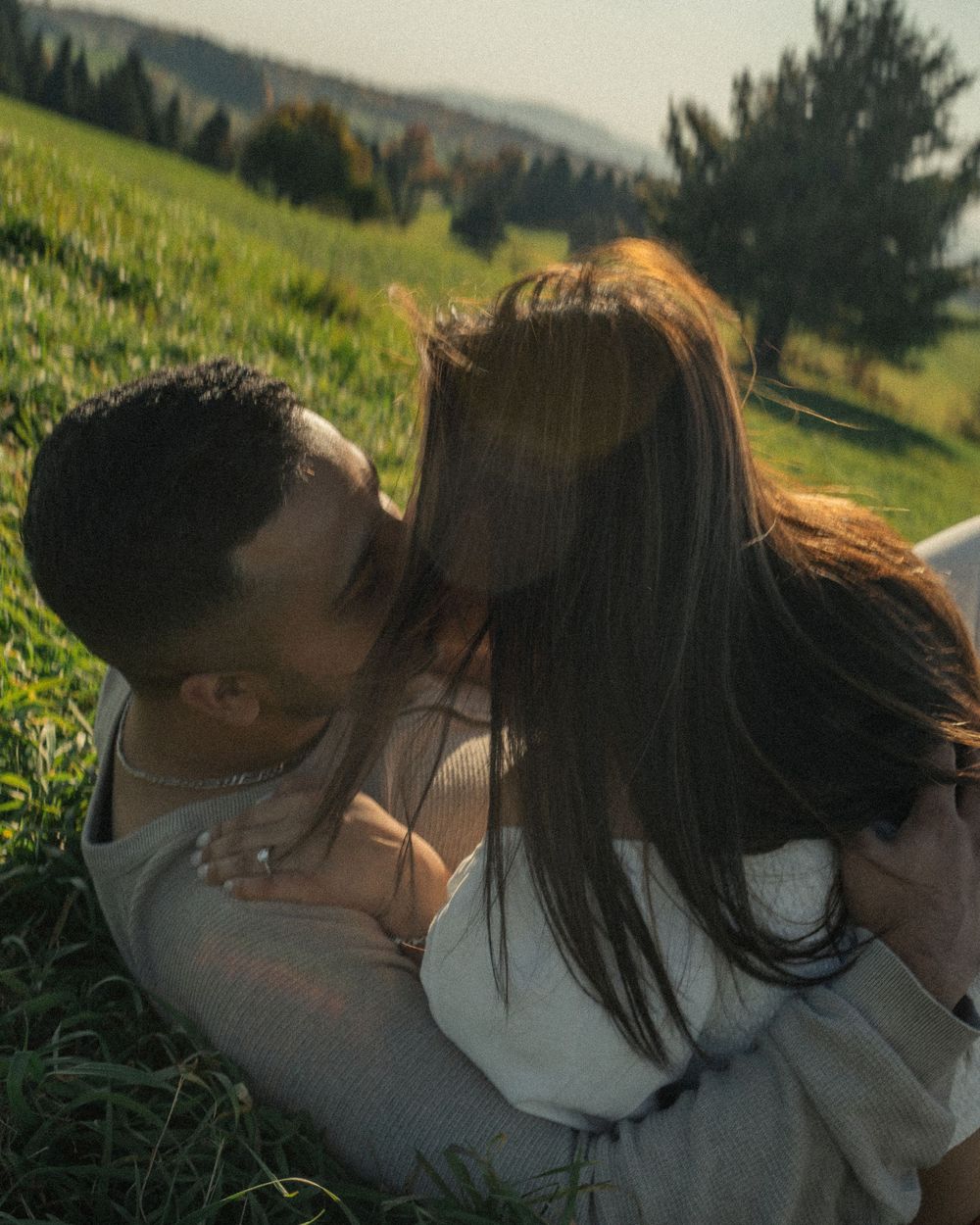 A romantic couple lying together in a sunlit grassy meadow with mountains in the background.