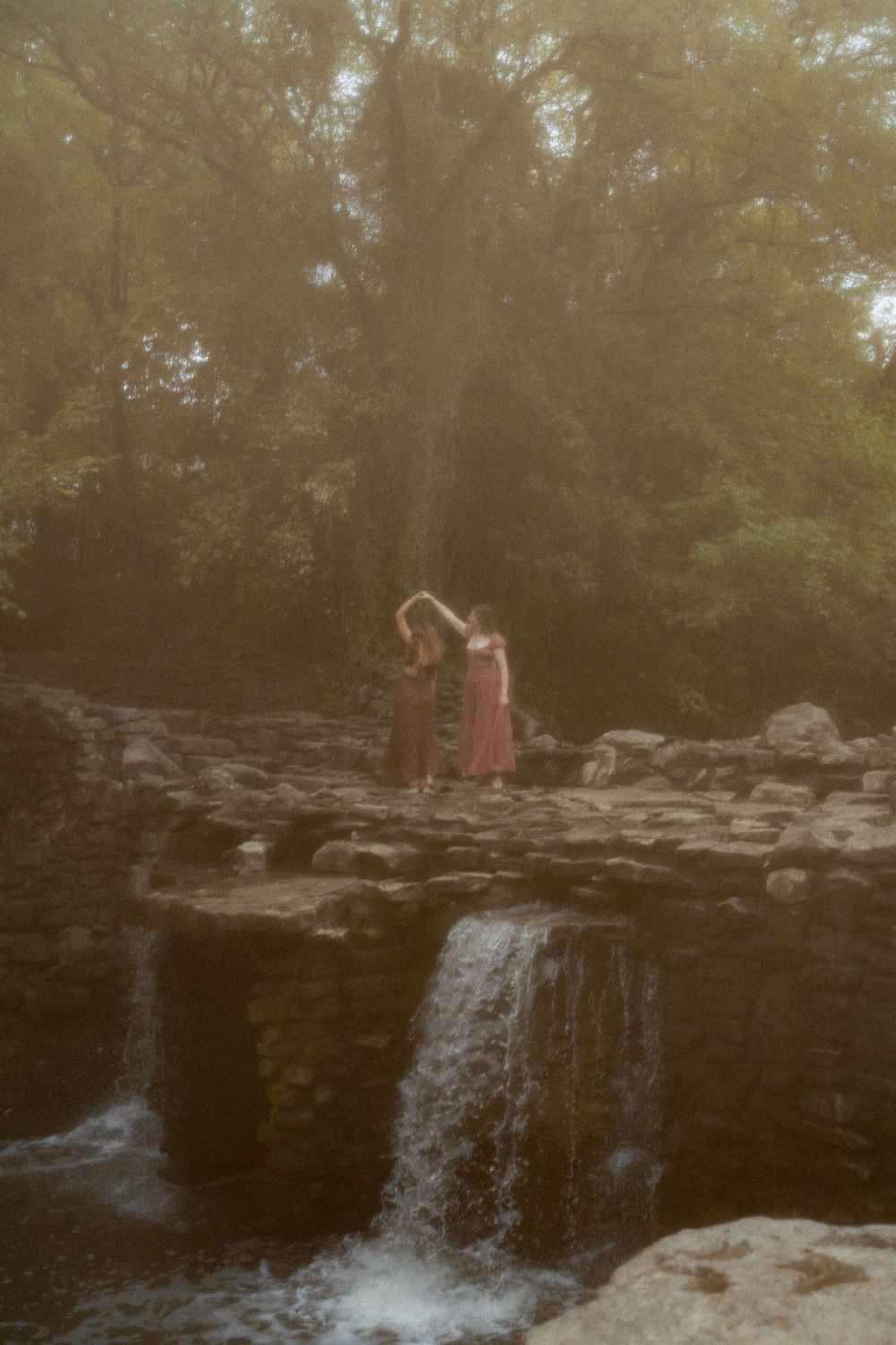 Figure in a red dress standing on rocky ledge near a waterfall at dusk.