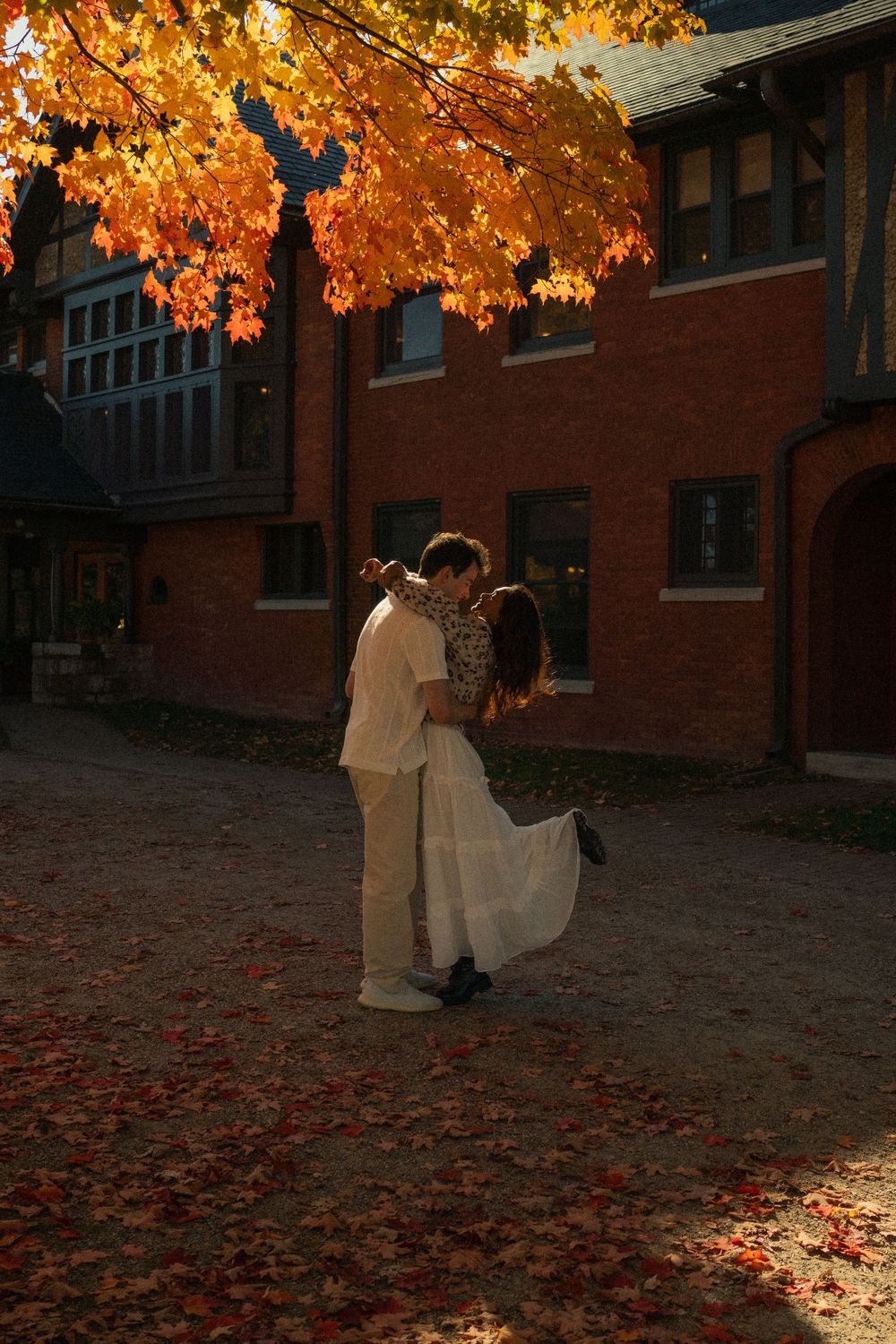 A romantic autumn scene with falling leaves as a couple embraces outside a brick building.