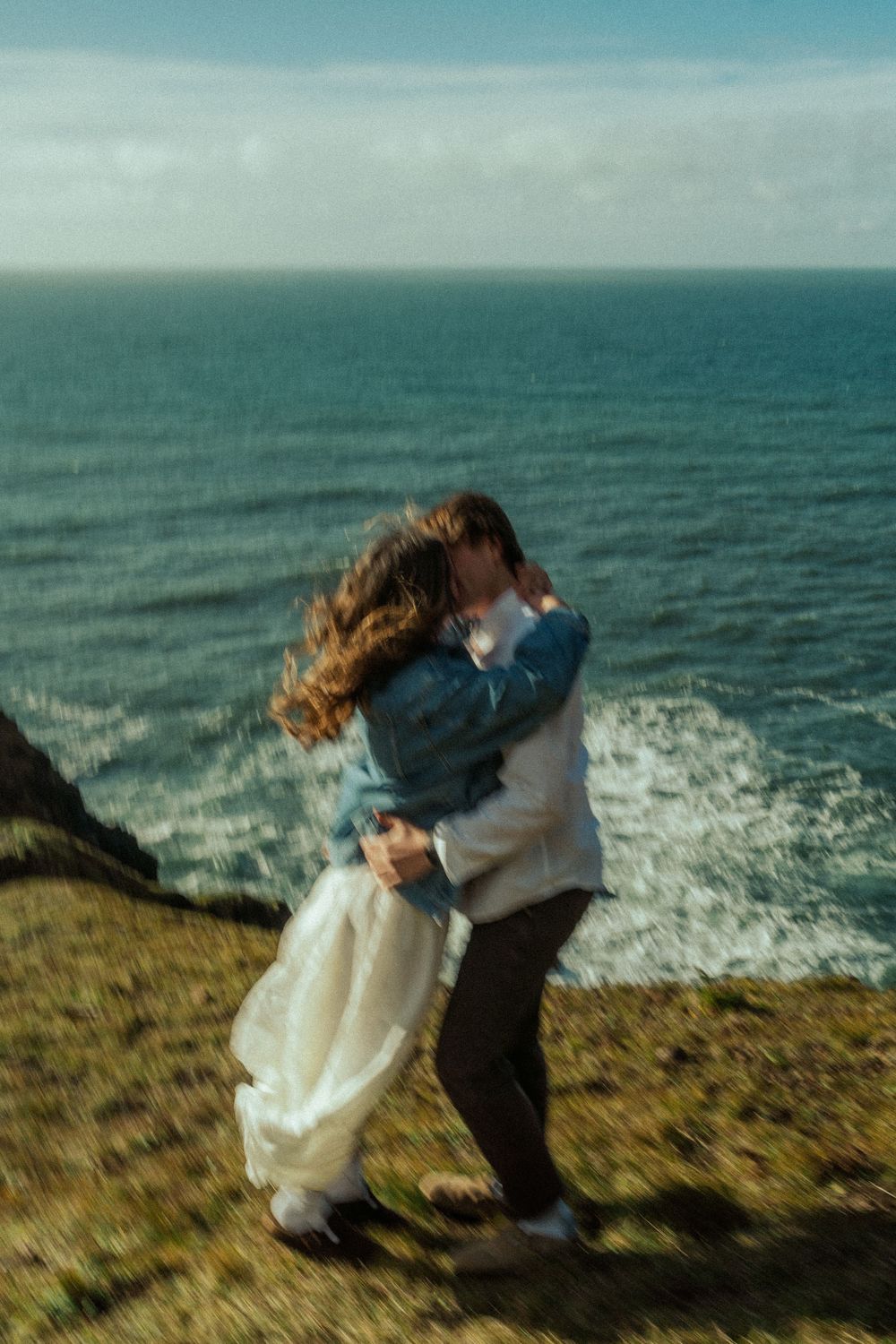 Couple embracing on coastal cliff edge with ocean waves crashing below.