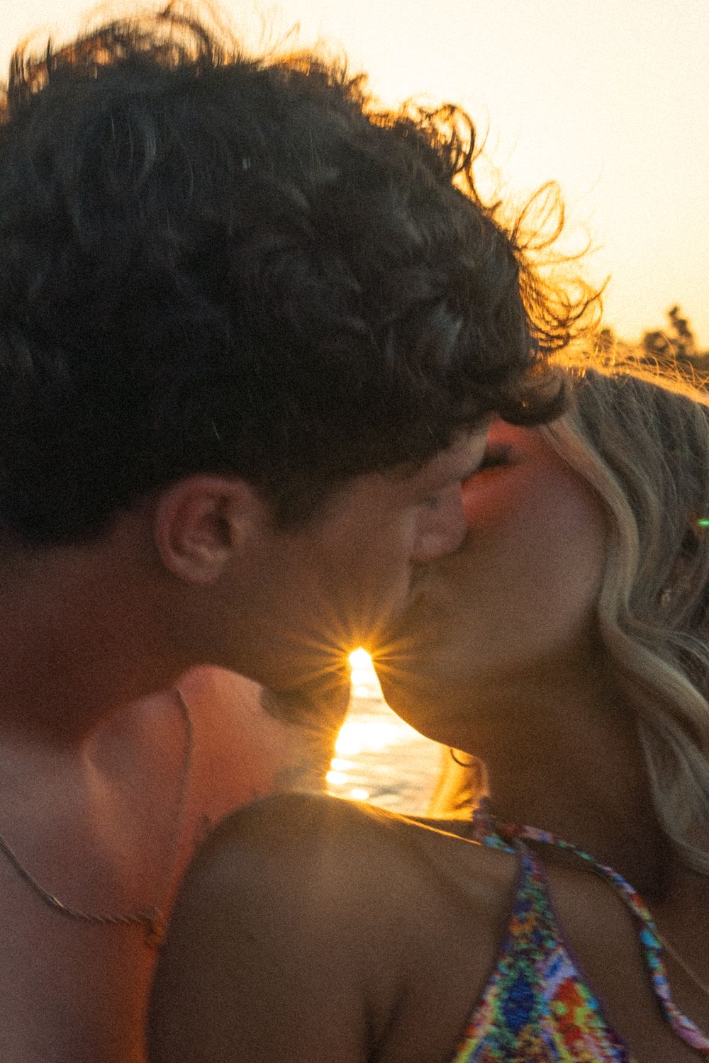 A couple sharing an intimate kiss at sunset with sun flare between them.