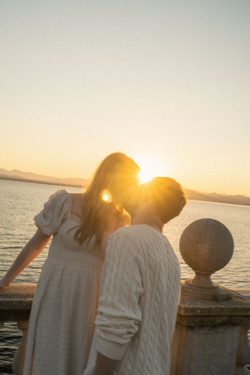 A couple in white outfits stands close together against an ocean sunset backdrop.