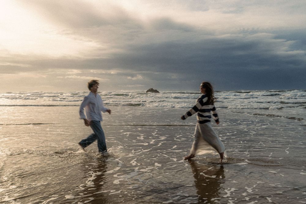 Two figures playfully chase each other along a misty beach at sunset with waves crashing in the background.