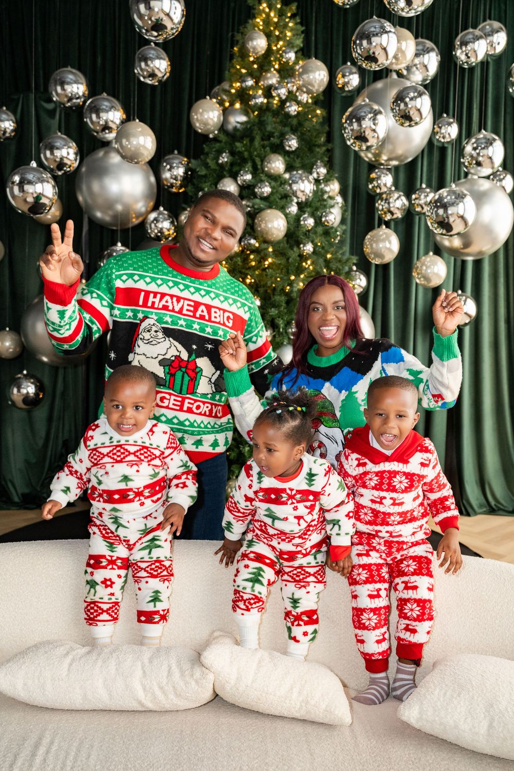 Family in matching Christmas pajamas poses by decorated tree with silver ornaments and green curtains.