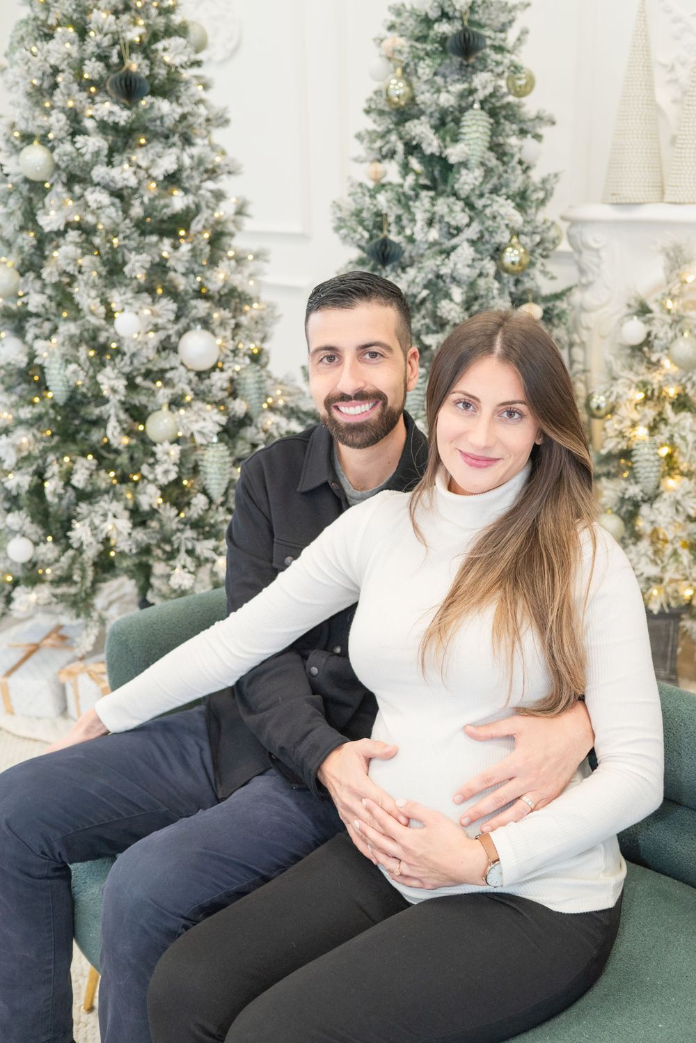 A couple poses on a green chair in front of Christmas trees, showcasing an expectant mother's baby bump.