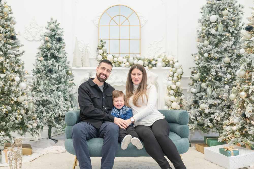 A family of three sits on a teal couch surrounded by flocked Christmas trees and holiday decor.
