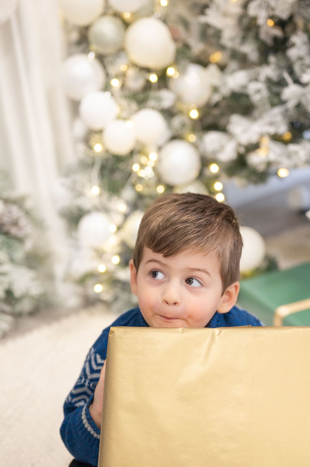 A child peeks over a large gold gift box near a Christmas tree decorated with white ornaments.