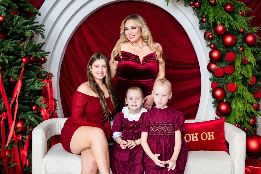 Family Christmas photo with woman and three girls in red dresses posed on white sofa with festive holiday decorations.