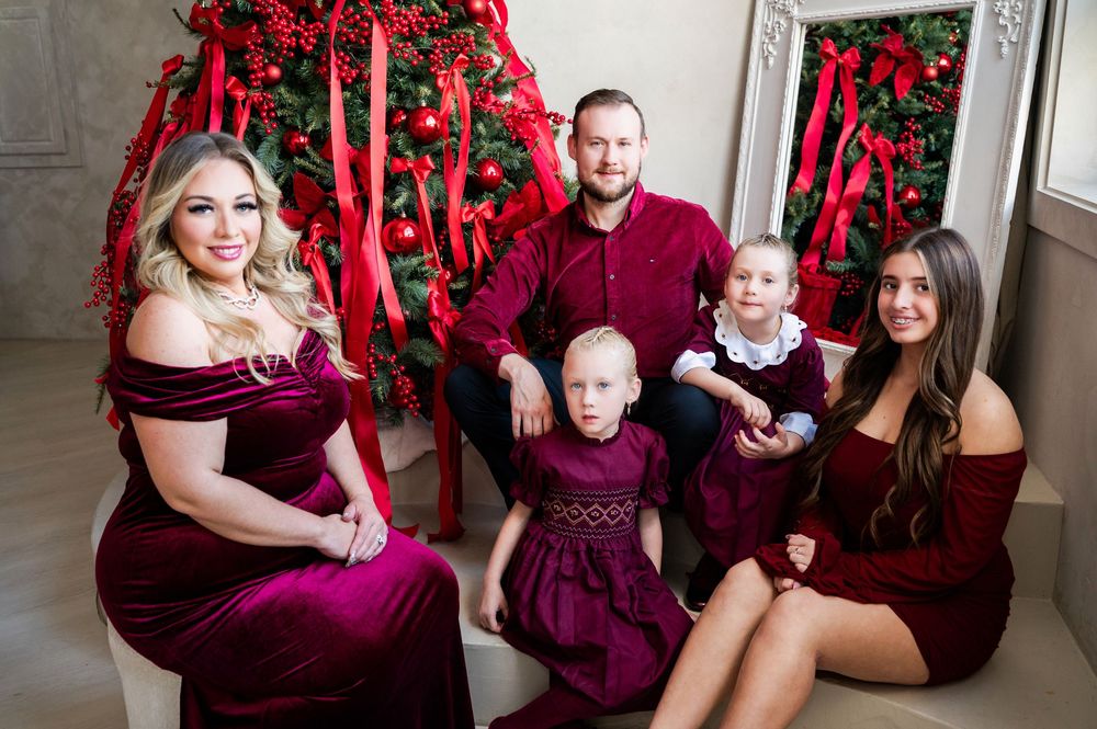 Family of five posing in coordinating burgundy outfits for a festive Christmas photo in front of a decorated holiday tree.