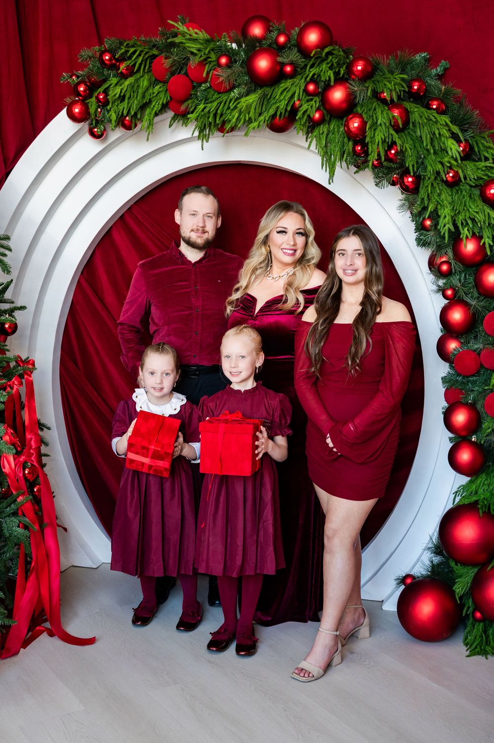 Family of five posing in festive red outfits under a Christmas wreath arch with red ornaments and holiday decor.