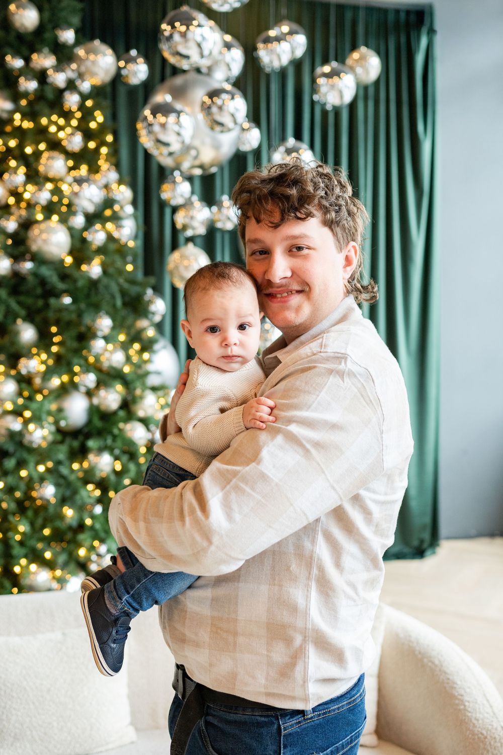 Father holding baby in front of decorated Christmas tree with silver ornaments and green curtains.