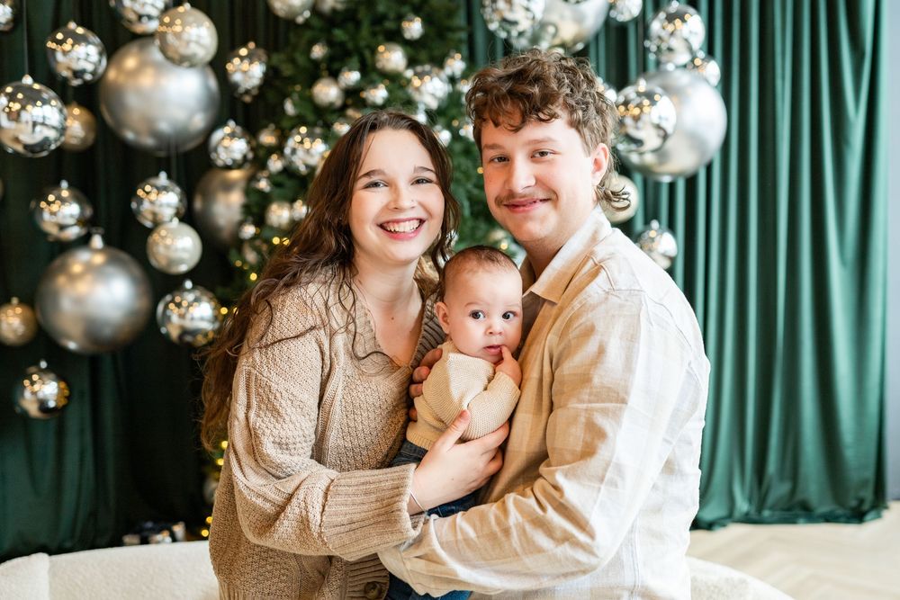 Young family smiling with newborn baby in front of decorated Christmas tree with silver ornaments.