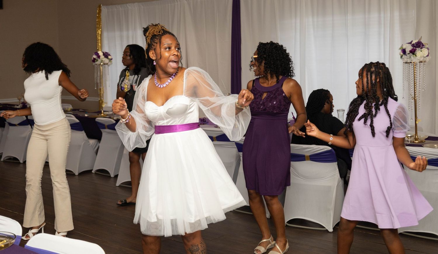 Bride in white dress dancing joyfully with guests at a purple and white wedding reception.