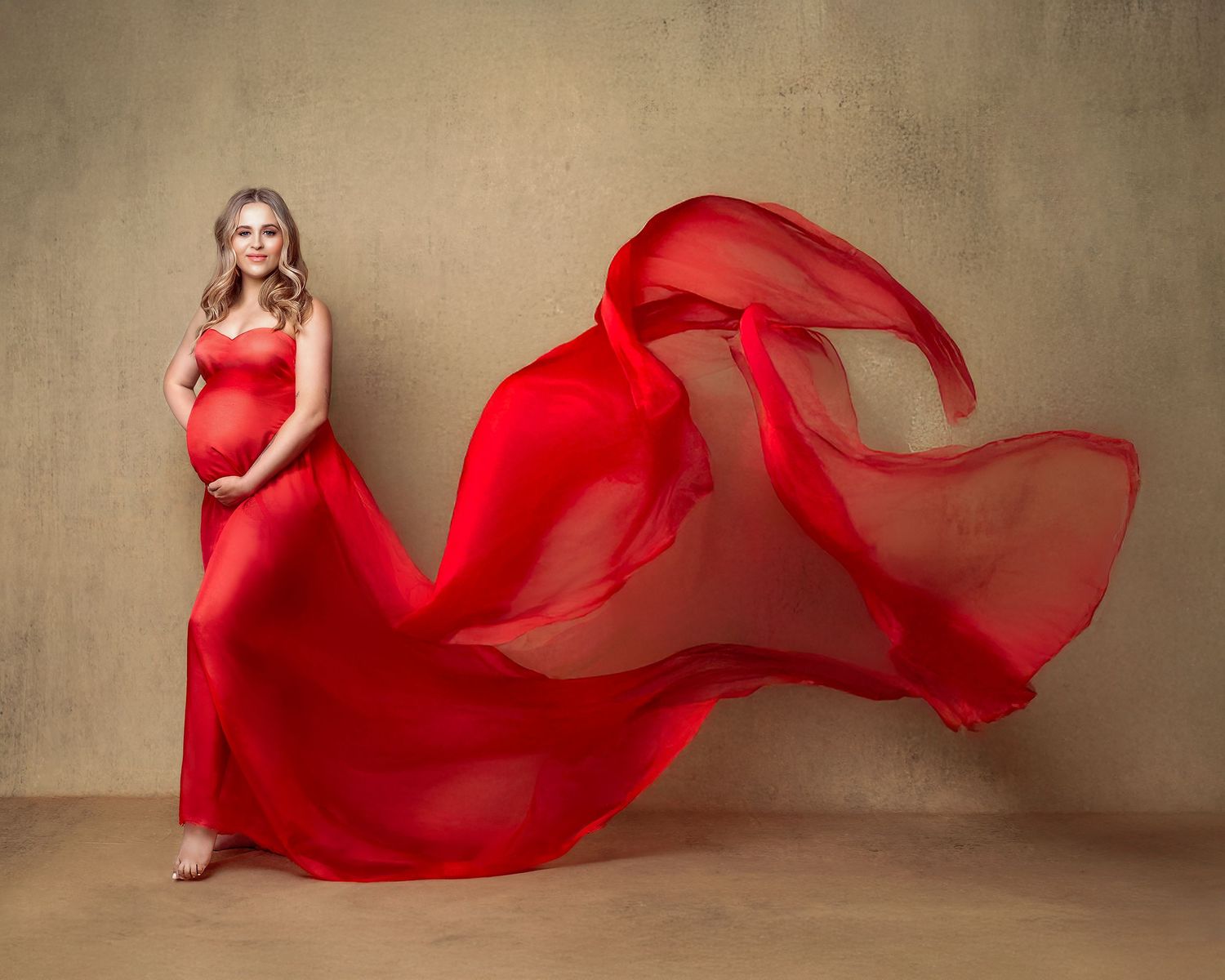 Elegant woman in a flowing red chiffon scarf, against a gold backdrop, celebrating her pregnancy with Caz Captures.