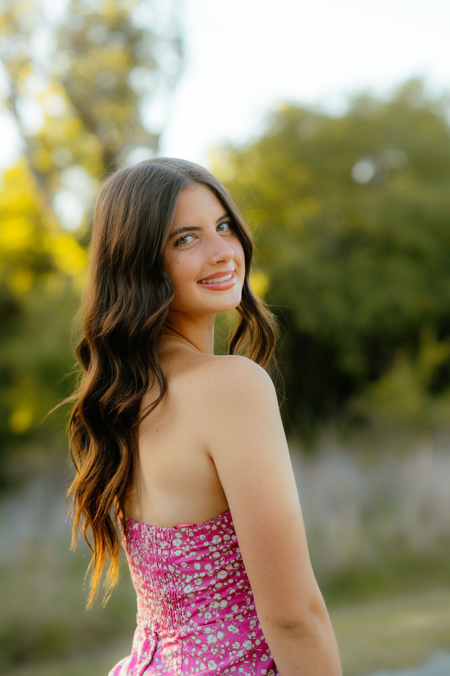 Senior girl in a floral dress smiling in tall grass during golden hour for Enid senior photos.