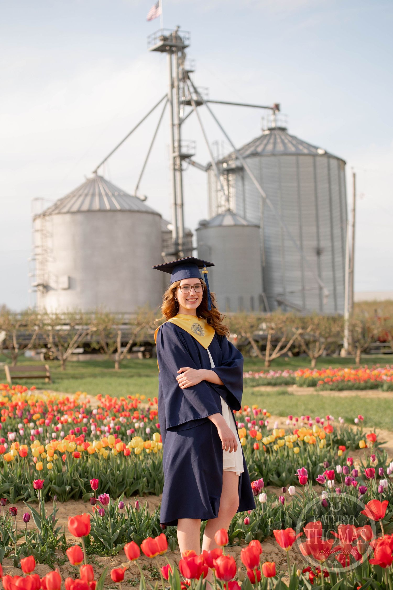 Graduate stands in colorful tulip field with large metal grain silos in background during spring photo session.