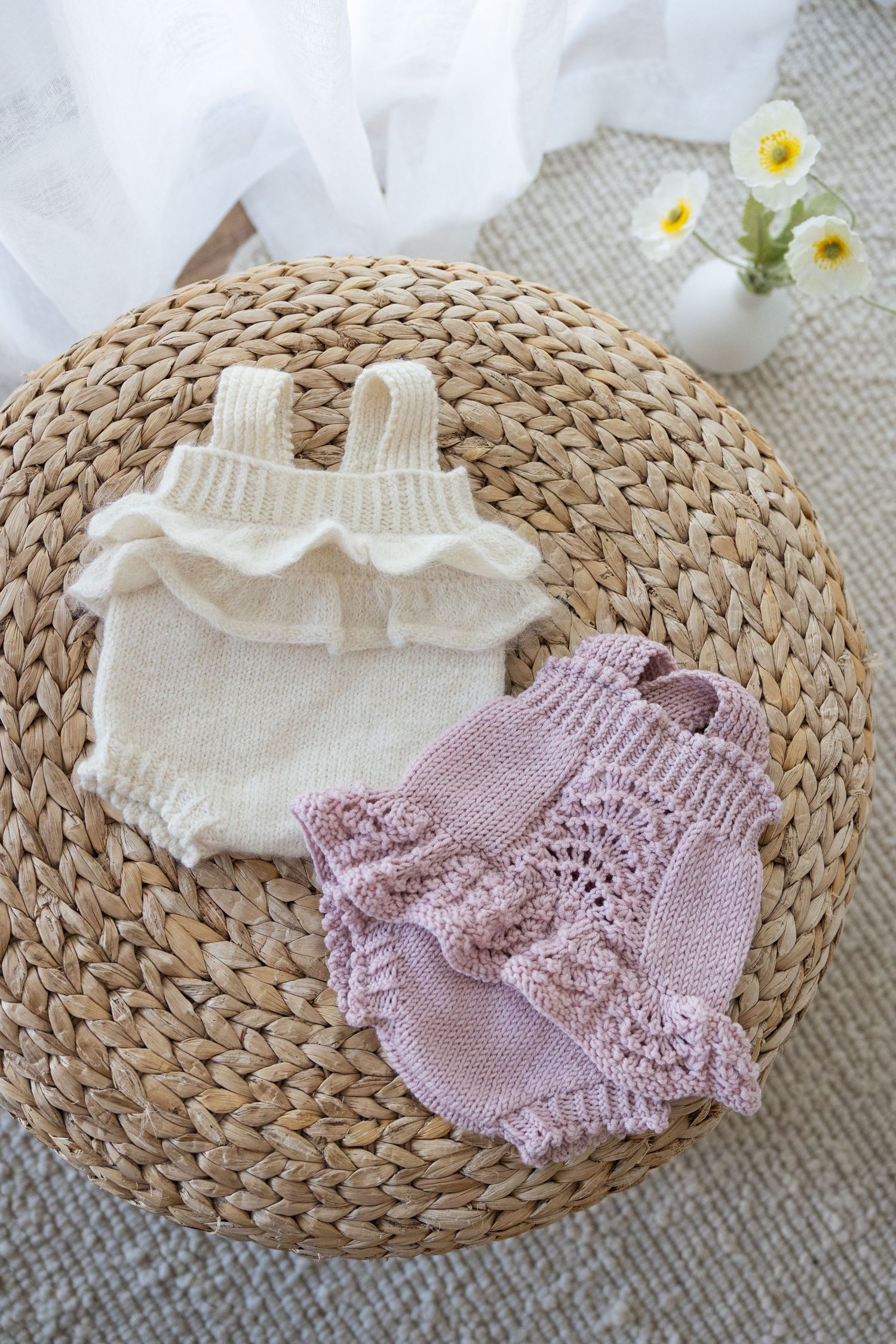 Pink knit baby romper with buttons displayed on a round woven straw mat with white daisies in the background.