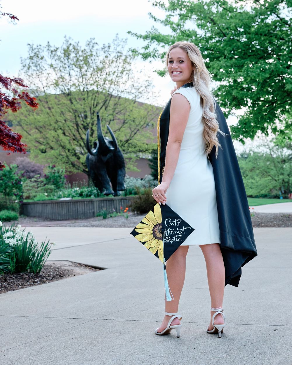 Stylish graduate poses with her decorated cap and gown on a campus path, with a sculpture in the background; professional graduation photo.