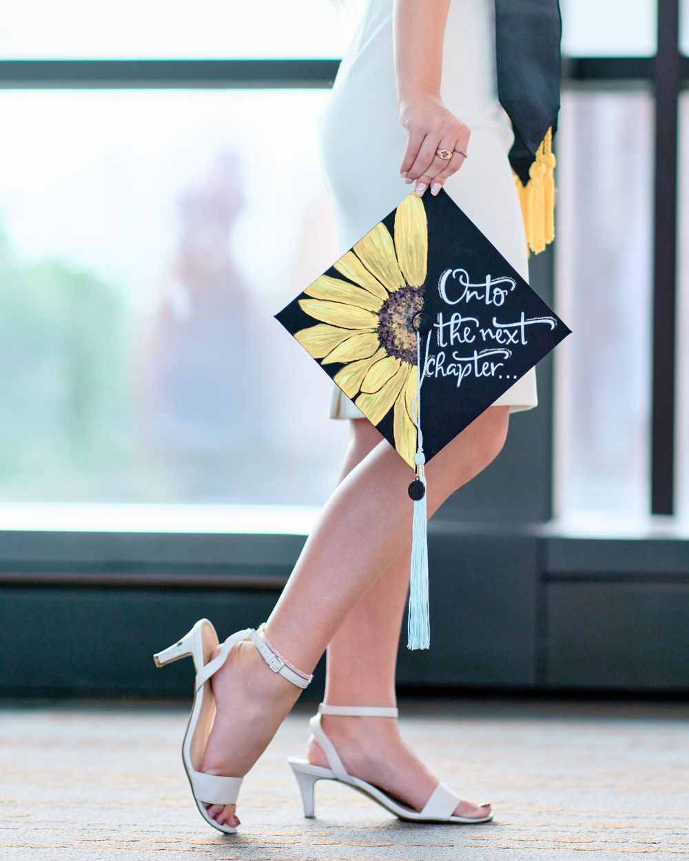 Close-up of a graduate holding a sunflower-decorated cap reading 'Onto the next chapter...'; creative graduation portrait detail.