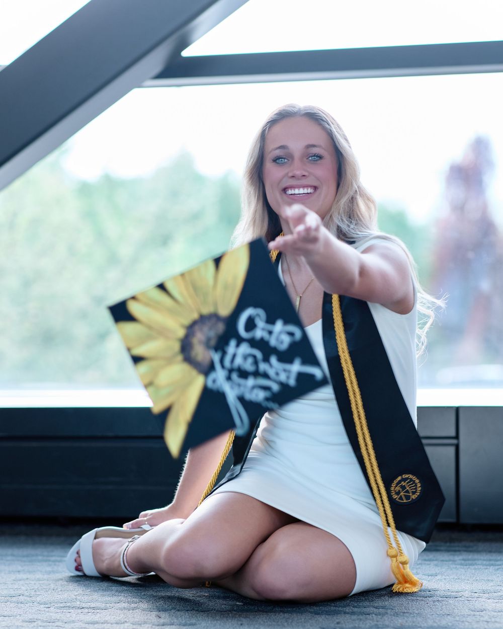 Smiling graduate in a white dress sits by a bright window, holding out her sunflower-decorated cap; indoor senior portrait photography.