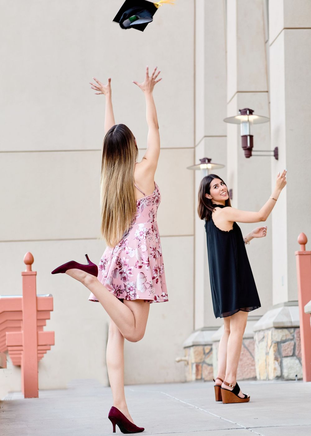 Joyful graduate in a pink dress tosses her cap in the air while a friend watches, celebrating outdoors; candid graduation photography.
