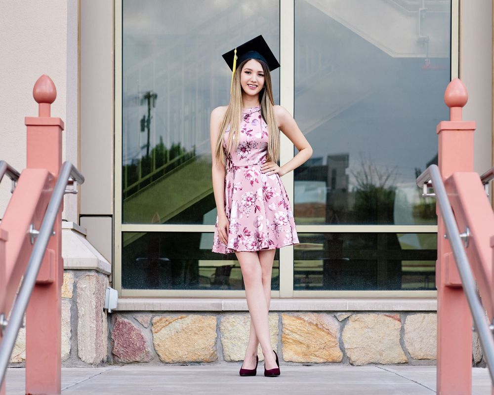Outdoor graduation portrait of a smiling young woman wearing a cap and dress, standing confidently by a building; natural light photography.
