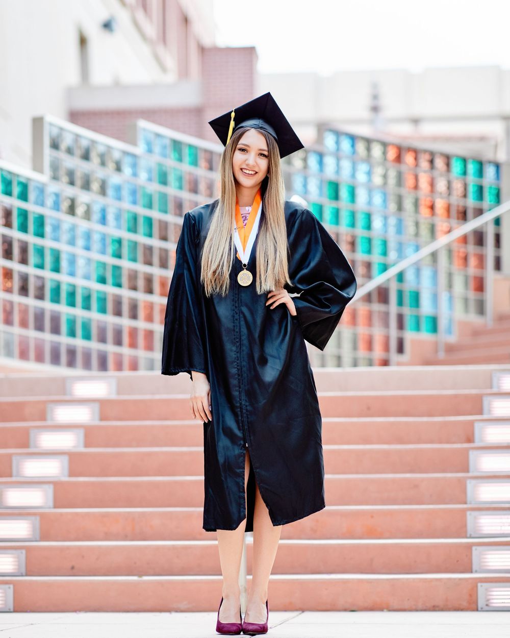 Joyful graduate in cap and gown, posing confidently on outdoor steps; professional graduation portrait photography.