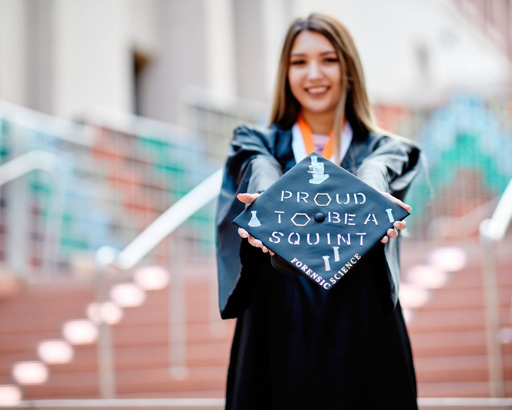 Graduate proudly displays her decorated 'Forensic Science' cap towards the camera; creative outdoor graduation portrait.