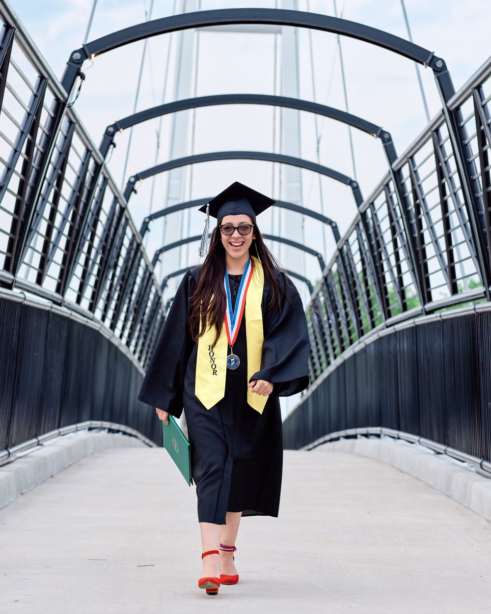 Smiling graduate in cap and gown walks confidently across a modern bridge, holding her diploma; outdoor graduation portrait photography.