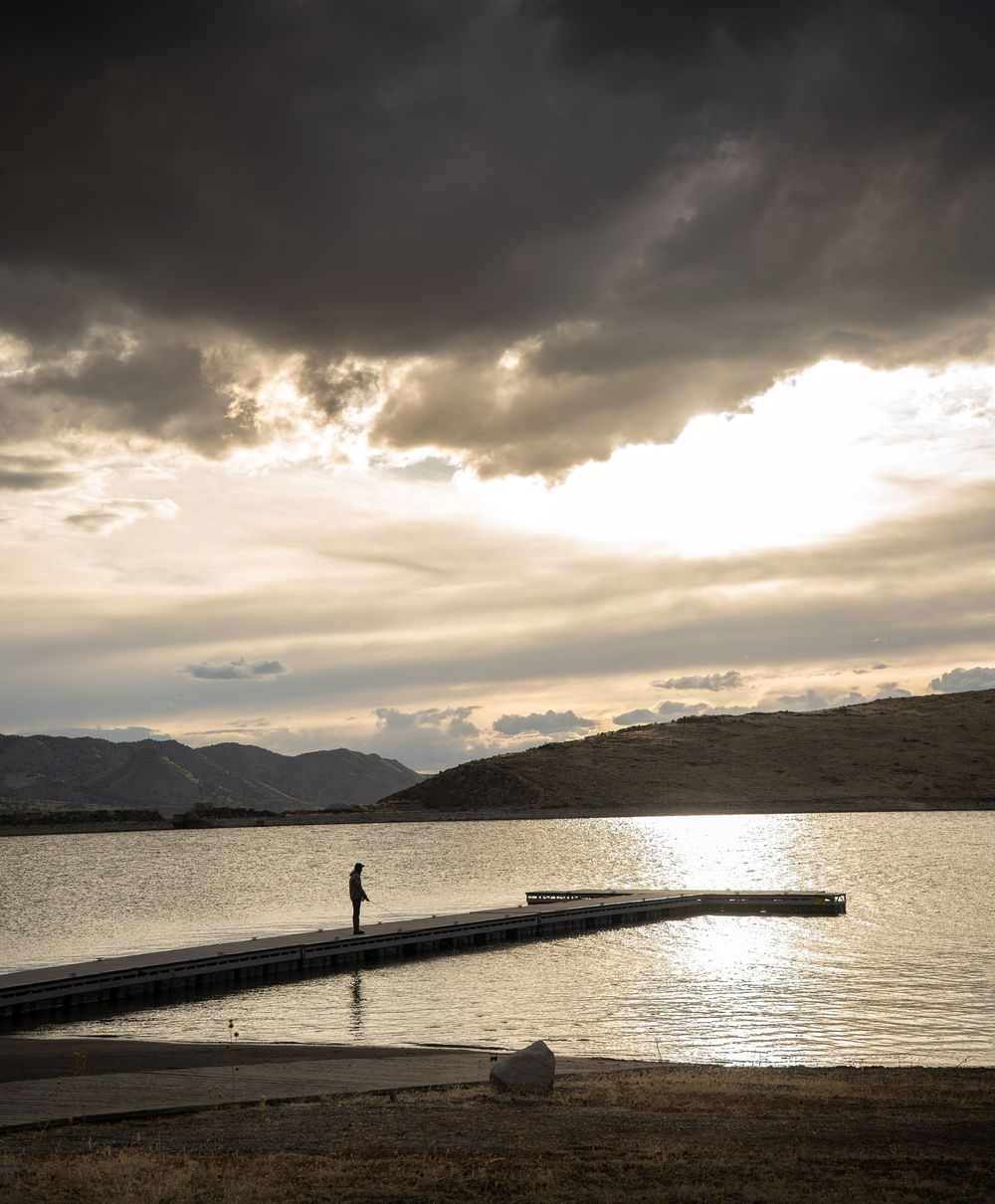 A dramatic panoramic seascape with dark storm clouds and a silhouetted figure standing on a pier at sunset.