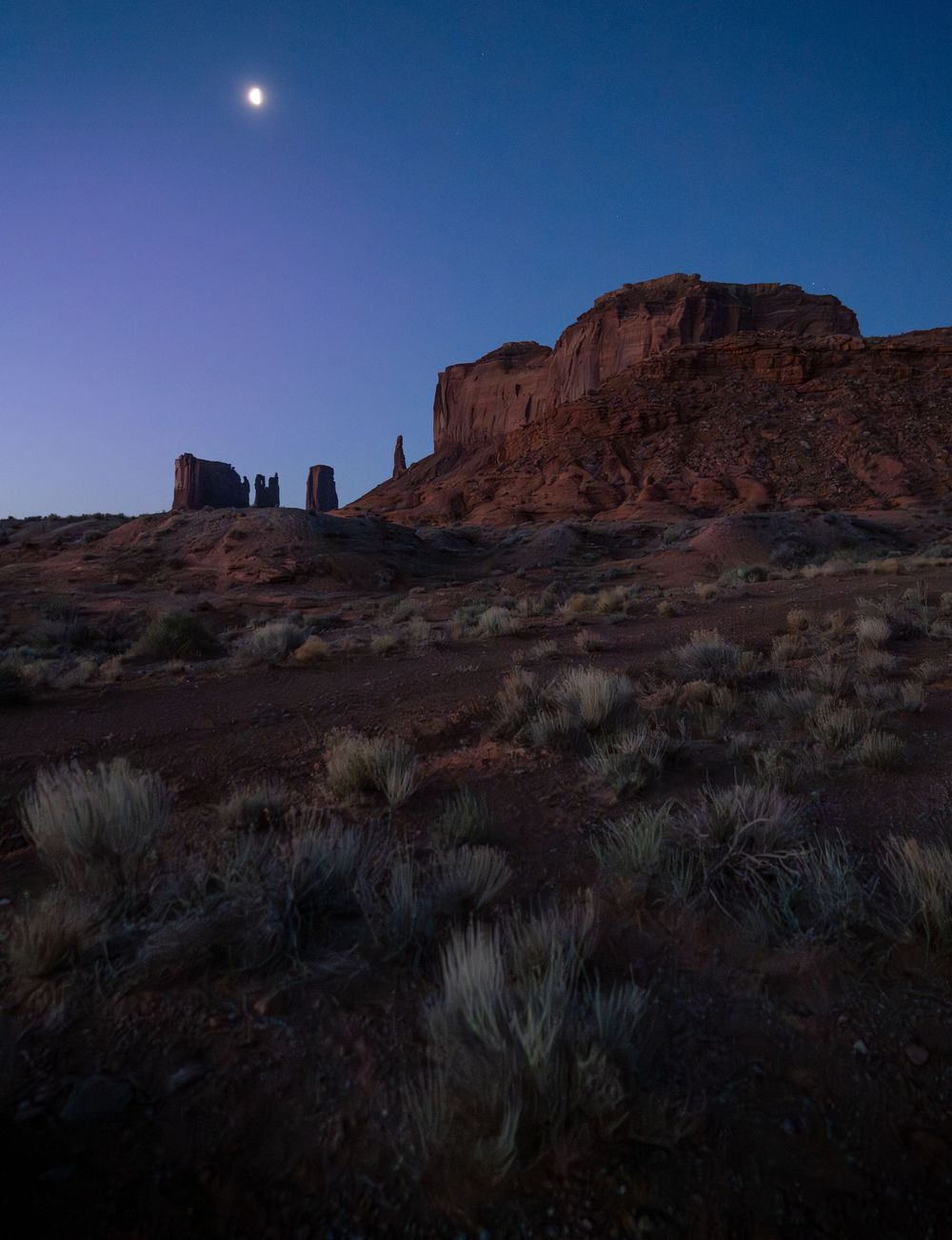 Moonlit landscape at Monument Valley shows red rock formations silhouetted against darkening twilight sky with desert brush.