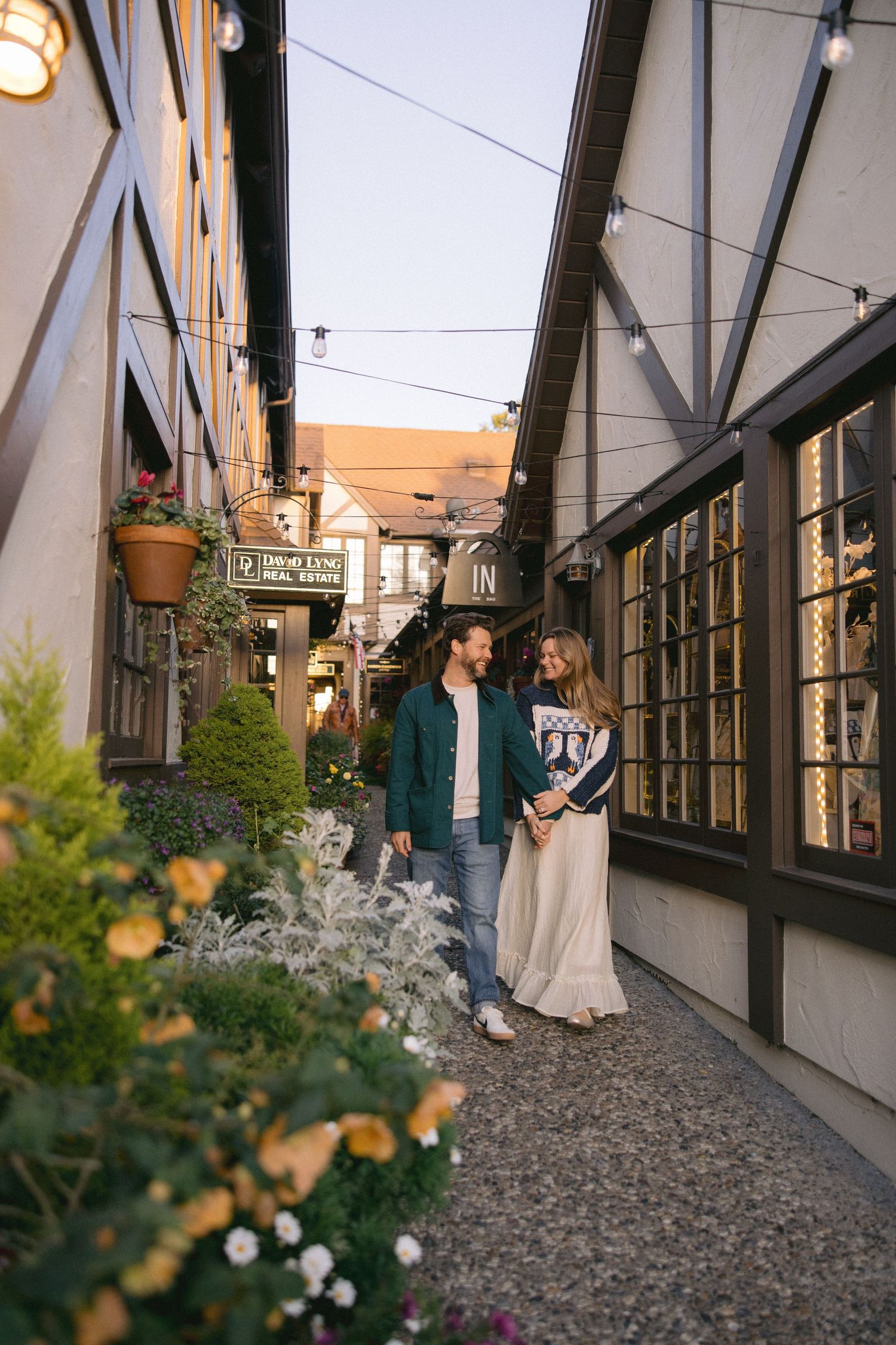 Two people walk down a charming cobblestone alley lined with flowers and storefronts at dusk.