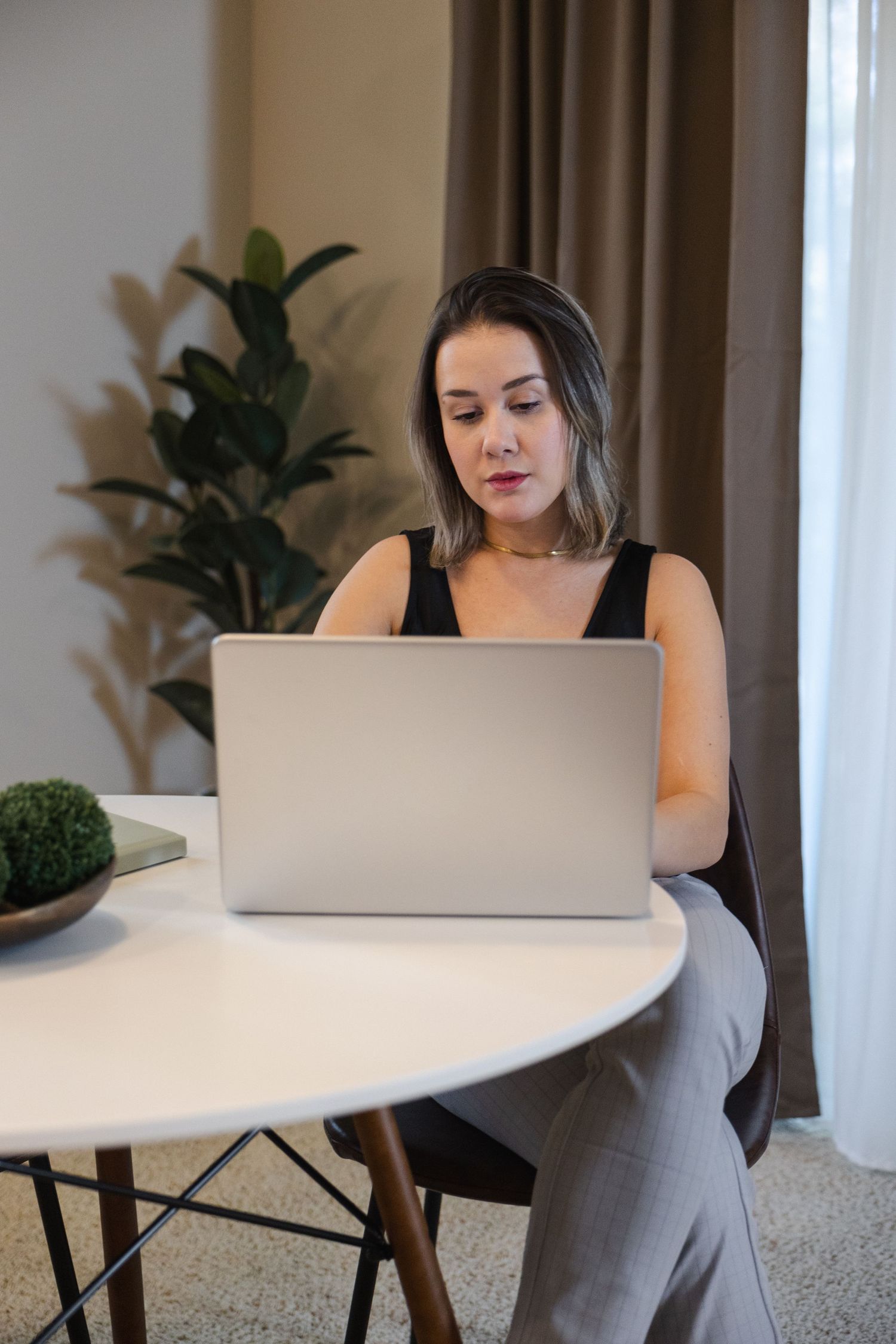 Someone working at home on a laptop at a round white table with a house plant in the background.