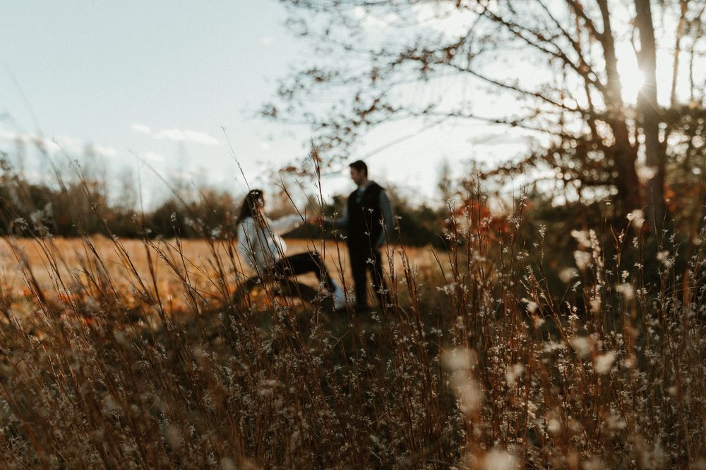 A couple stands in a misty field during golden hour with autumn foliage in the background.