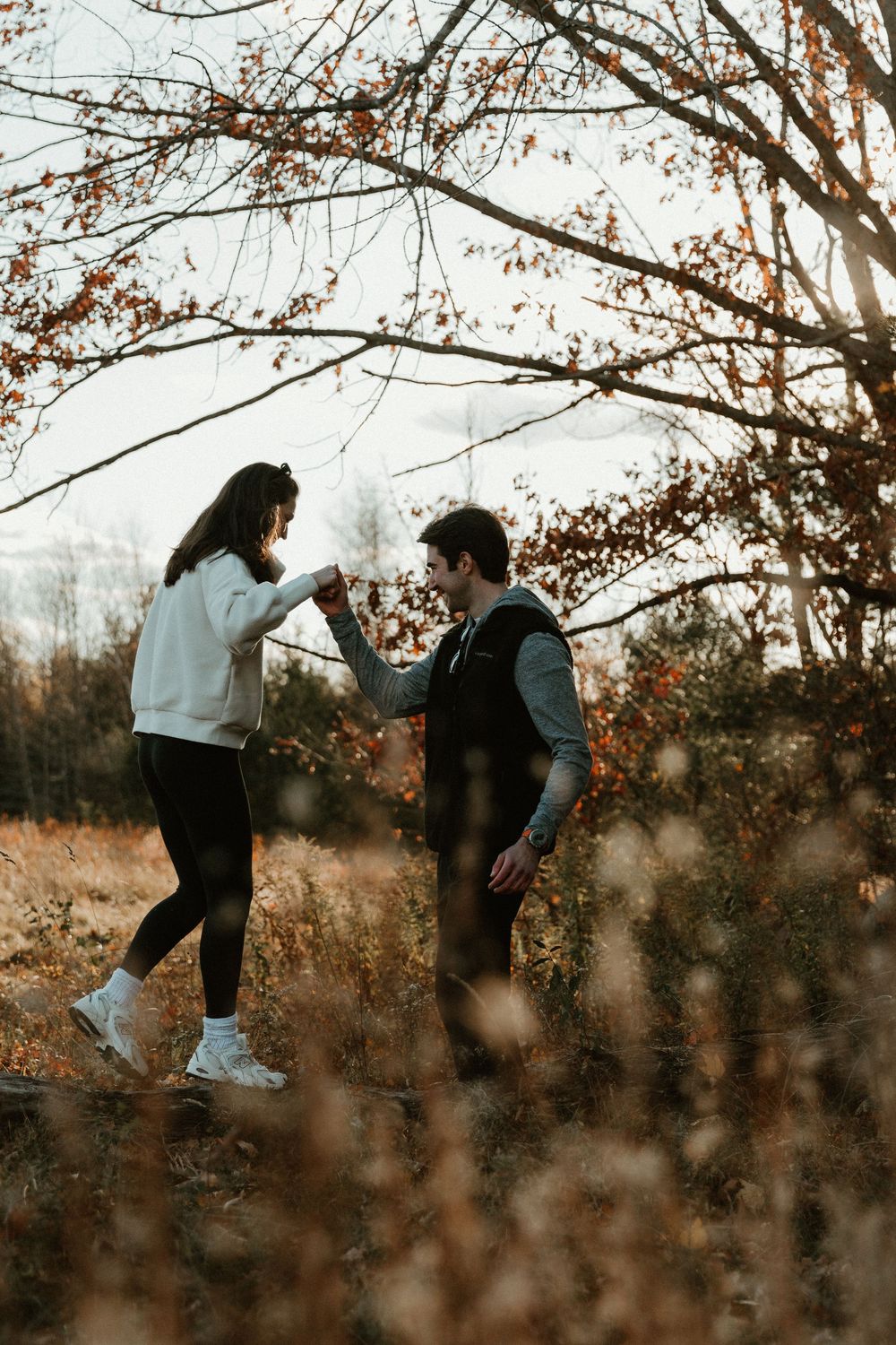 Two people share a playful moment on a fallen tree branch in an autumn field.