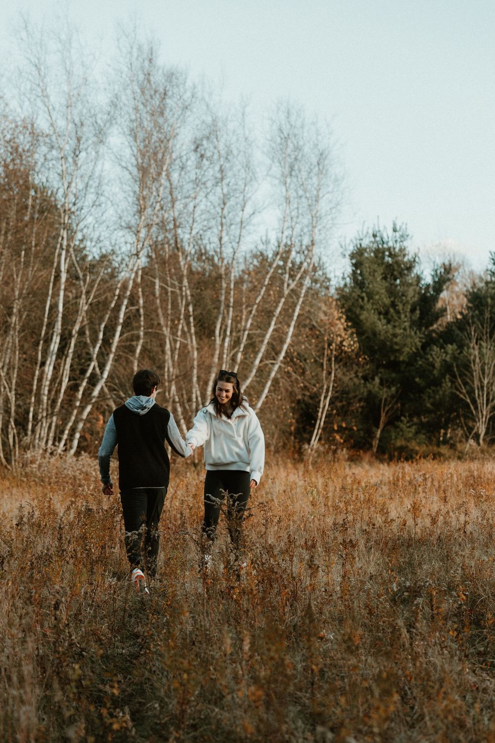 A couple walks together through a field of tall grass with white birch trees in the background during autumn.