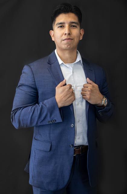 A young man in a blue suit and white dress shirt adjusts his lapels against a dark background.