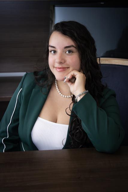 Young woman in green blazer and pearl necklace smiling while seated at a wooden table.