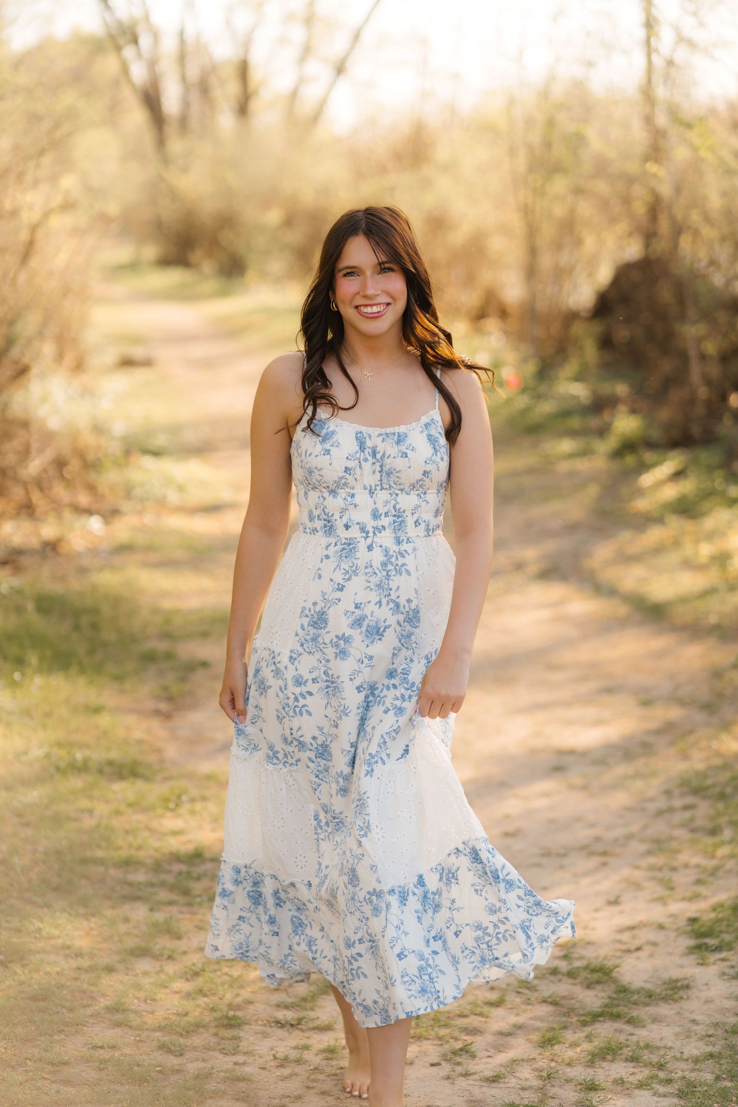 A flowy white and blue floral midi dress is worn on a sunny dirt path among trees in a dreamy outdoor setting.