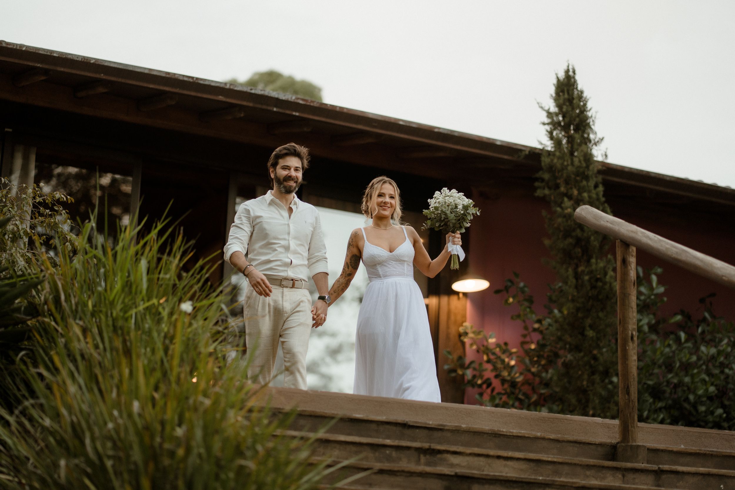 A couple descends wooden stairs at their outdoor wedding venue during sunset.